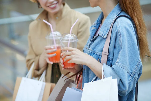 Close-up of positive ladies with shopping bags drinking sweet cocktails and chatting in mall