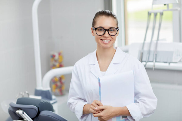 Waist up portrait of young female doctor wearing glasses looking at camera while posing in office, copy space