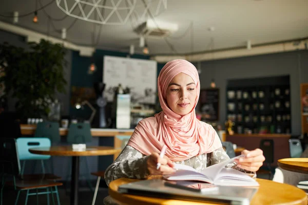 严肃的年轻穆斯林妇女坐在咖啡馆的桌子旁 看书和做笔记 — 图库照片