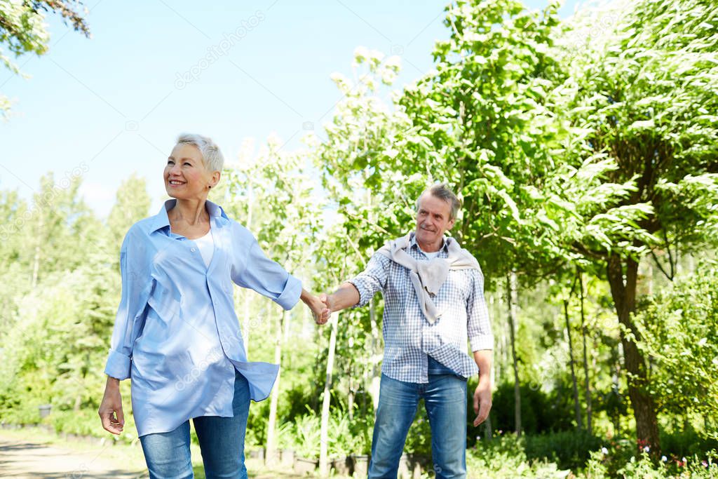 Portrait of happy senior woman leading husband by hand while enjoying walk in Summer park, copy space