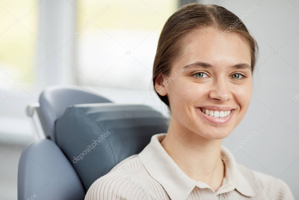 Head and shoulders portrait of pretty young woman smiling at camera and showing perfect white teeth while sitting in dental chair, copy space