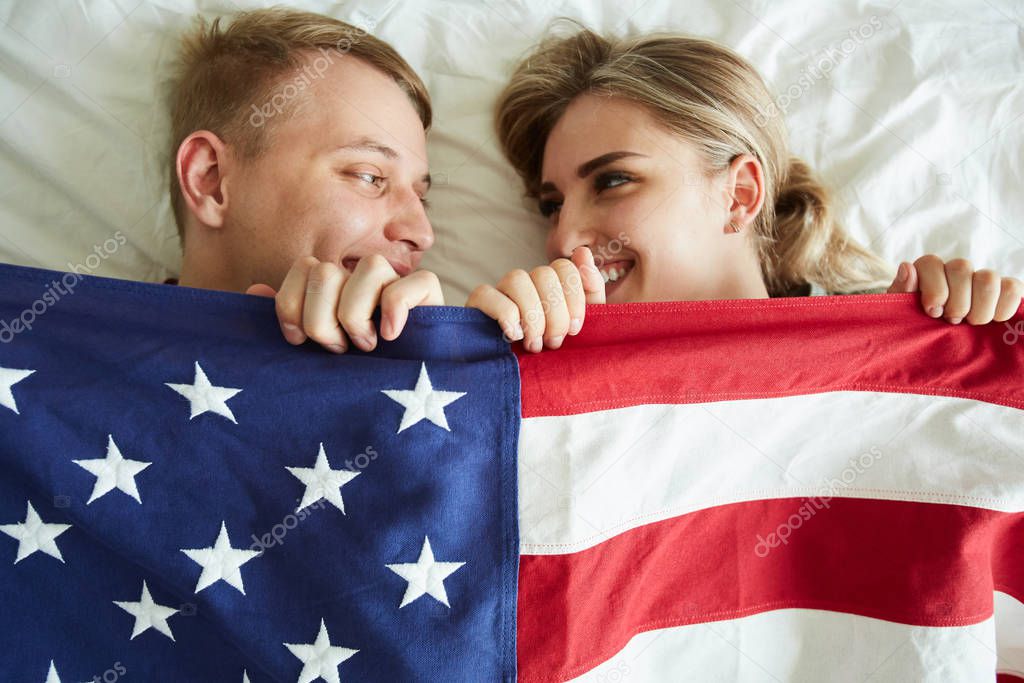 Happy young dates covering with American flag while lying on bed in front of each other
