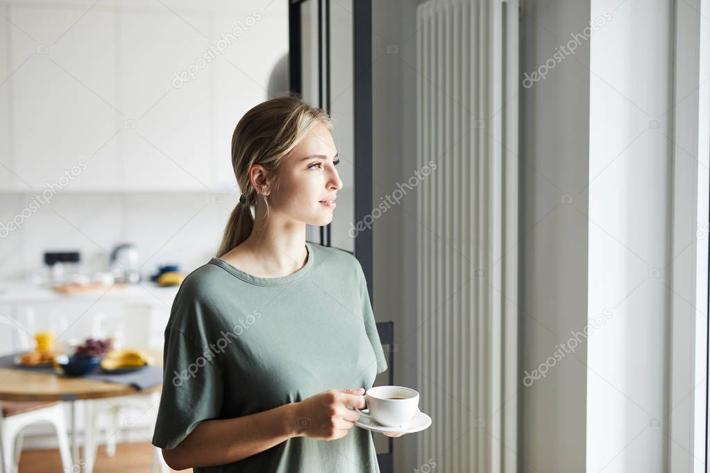 Young casual woman with cup of coffee looking through window while staying at home on weekend