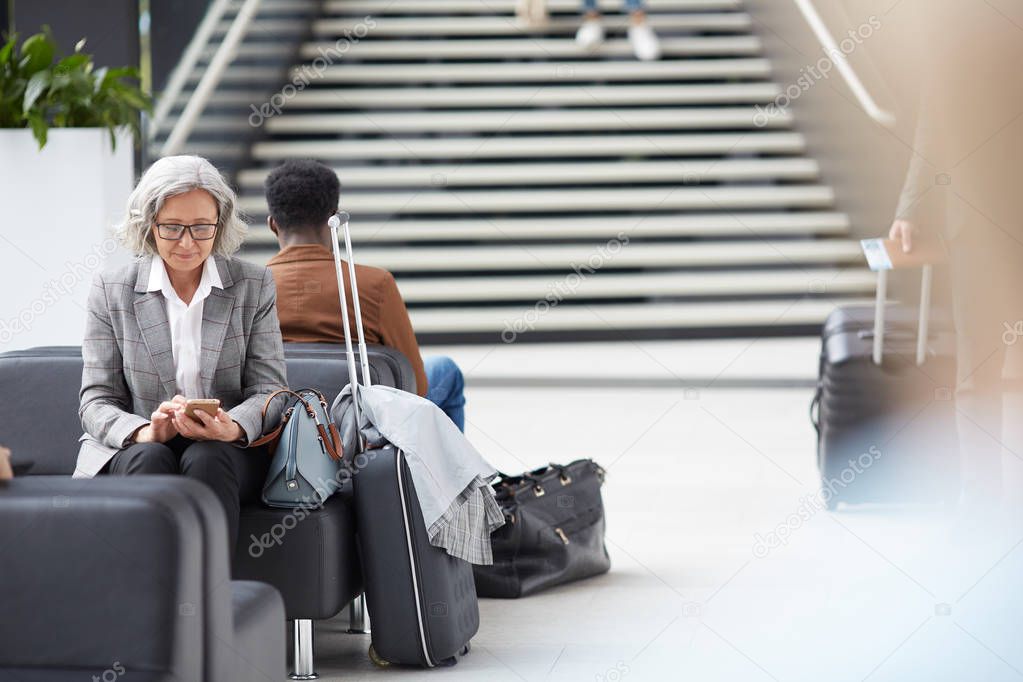 Content elderly Asian lady with gray hair sitting on black leather sofa in airport and browsing internet on smartphone