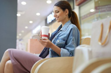 Alışveriş merkezinde kahve ile chilling ve onun akıllı telefon haber beslemesi kaydırma genç çekici kız
