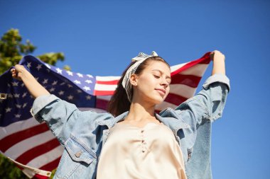 İçerik dalgın ücretsiz Amerikan kız şık başörtüsü ile rahat kıyafet açık havada ayakta ve Amerikan bayrağı tutan giyiyor