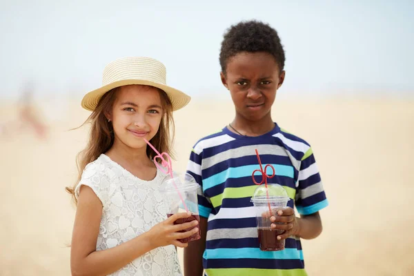 皱眉的非洲裔美国男孩和微笑美丽的女孩在沙滩上喝清爽果汁的肖像 — 图库照片