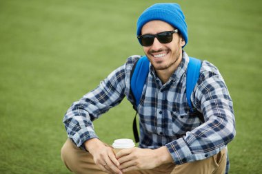 Modern backpacker portresi yeşil çim üzerinde oturan ve yalnız gezi sırasında kamera gülümseyerek, kopya alanı