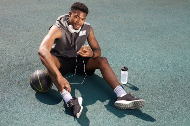Basketbol sahasında otururken ve akıllı telefon kullanırken kameraya bakan yakışıklı Afrikalı-Amerikalı adamın arka planı yukarıda, kopya alanı
