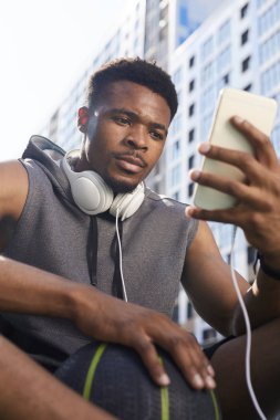Basketbol sahasında otururken akıllı telefon kullanan çağdaş Afrikalı-Amerikalı adamın düşük açılı portresi, kopya alanı