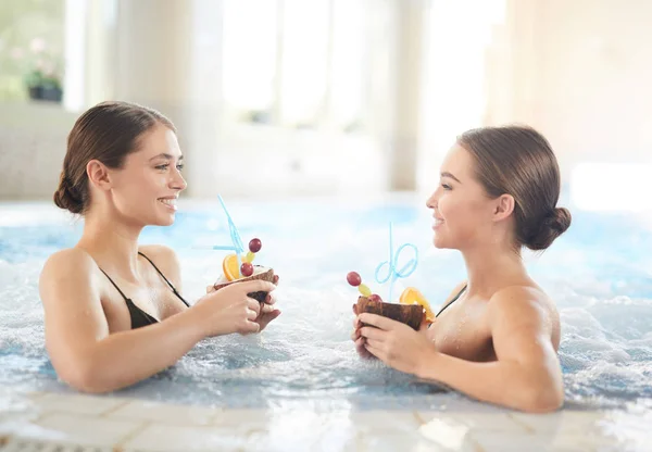Side view portrait of two beautiful young women smiling at camera while relaxing in hot tub enjoying spa, copy space