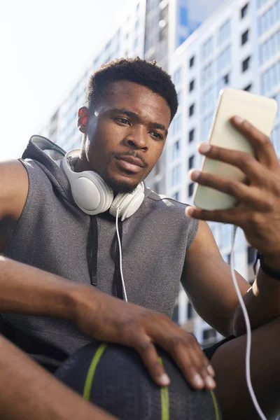 Basketbol sahasında otururken akıllı telefon kullanan çağdaş Afrikalı-Amerikalı adamın düşük açılı portresi, kopya alanı