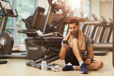 Yakışıklı adam spor salonunda yerde oturan ve akıllı telefon kullanarak tam uzunlukta portre, kopya alanı