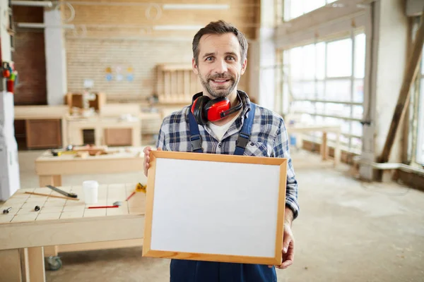 Waist up portrait of smiling mature carpenter holding blank frame and looking at camera, copy space