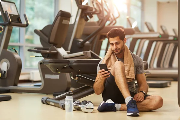 Yakışıklı adam spor salonunda yerde oturan ve akıllı telefon kullanarak tam uzunlukta portre, kopya alanı