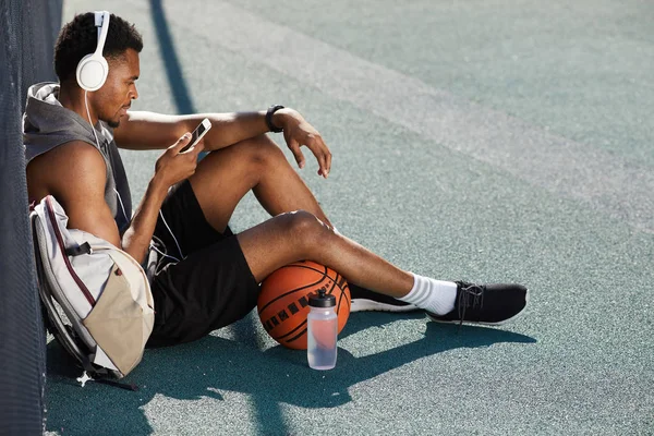 Basketbol sahası açık havada otururken kulaklık takan ve akıllı telefon kullanan çağdaş Afrikalı-Amerikalı adamın yüksek açılı portresi, kopya alanı