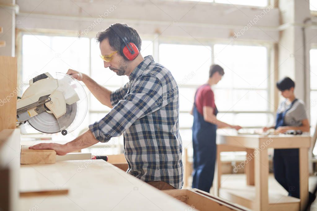 Side view portrait of mature carpenter sawing wood in industrial workshop, copy space
