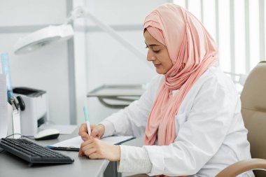 Orta Doğulu genç bir kadının portresi başörtüsü takıyor. Klinikte hemşire olarak çalışıyor ve hasta formlarını dolduruyor.