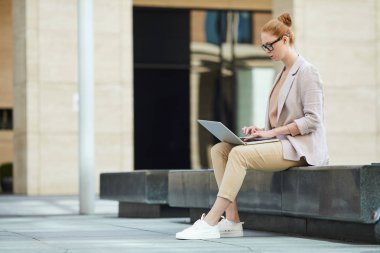 İşletme projesi için kablosuz ağ ve kopya temposu kullanırken dışarıda dizüstü bilgisayar kullanan kızıl saçlı genç kadının yan görünüm portresi