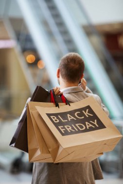 Alışveriş merkezinde yürüyen merdivene karşı konuşurken, üzerinde Kara Cuma yazıları olan alışveriş torbaları tutan modaya uygun bir adamın arka plan portresi, kopyalama alanı
