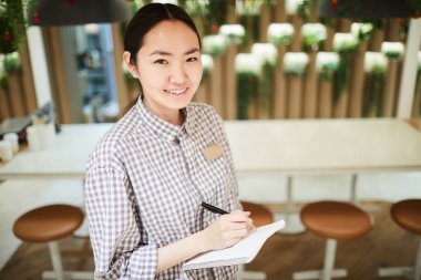 High angle portrait of smiling Asian waitress looking at camera while taking order in cafe, copy space