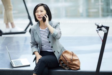 Modern Asyalı kadının soğuk tonlu portresi. Akıllı telefonuyla konuşuyor. Açık beton bankta oturuyor.