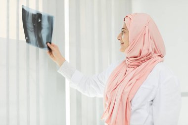 Doktor olarak çalışan Arap kadının yan görüş portresi. Pencere önünde bacak eklemlerinin röntgenini tutuyor, boşluğu kopyalıyor.