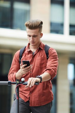 Şehir ortamında elektrikli scooter sürerken akıllı telefonu kontrol eden yakışıklı bir gencin belden yukarı portresi.