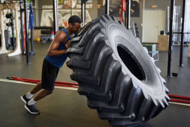 Modern spor salonunda spor yaparken lastiği ters çeviren kaslı Afrikalı adamın tam boy kenar görüntüsü.