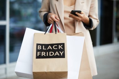 Kara Cuma 'yla alışveriş torbaları taşıyan şık bir genç kadının orta sınıf portresi ve alışveriş merkezinden ayrılırken mesajlaşması.