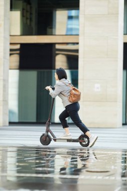 Full length side view of young Asian woman riding electric scooter fast in city street, copy space