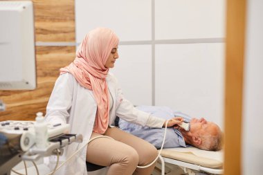 Tıbbi klinikteki yaşlı hastaya ultrason uygulayan genç Arap kadın doktorların yan görüş portresi.