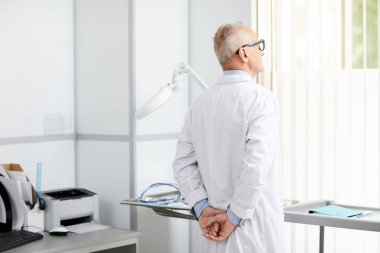 Modern kliniğin ofisinde dikilirken pencereye bakan düşünceli kıdemli doktorun portresi.