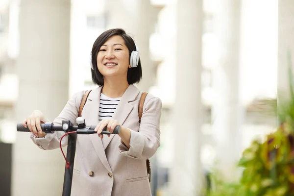 Waist up portrait of adult Asian woman riding electric scooter and smiling happily at camera in city street, copy space