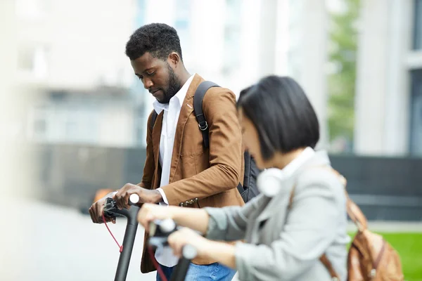 Side view portrait of two people riding electric scooters in city street, focus on African-American man in background, copy space