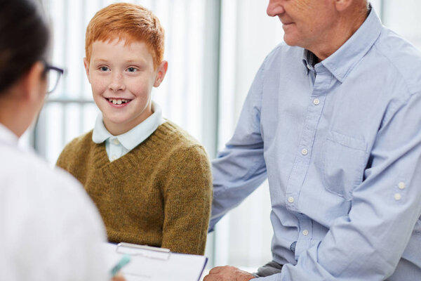 Portrait of smiling boy visiting doctor with father and looking at nurse filling patients form in waiting room, copy space