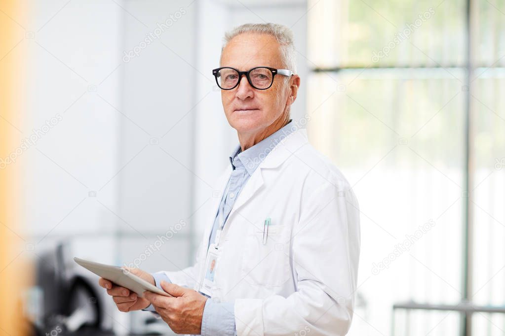 Waist up portrait of senior doctor looking at camera while using digital tablet standing in office of modern clinic, copy space