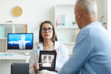 Portrait of serious female doctor holding x-ray image of lungs and chest while consulting senior patient in clinic, copy space clipart