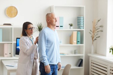 Yaşlı bir hastanın kalp ve nefes alışını dinleyen, klinikteki muayene sırasında steteskop takan kadın doktorların yan görüş portresi.