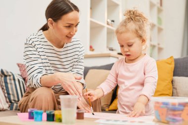 Young woman spending time with daughter at home teaching her to paint pictures, horizontal shot clipart