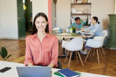 Pembe bluz giyen neşeli genç bir kadın ofis masasında oturmuş kameraya bakıp gülümsüyor ve iş arkadaşları arkasında çalışıyor.