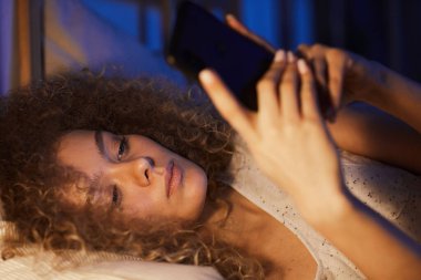 Geceleri yatakta akıllı telefon kullanan kıvırcık saçlı genç bir kadının portresini yakın çekin. Ilık lamba ışığı, fotokopi odası.