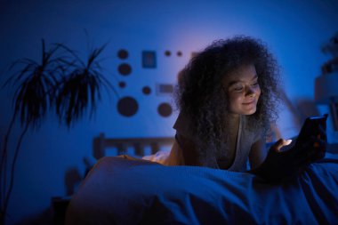 Akıllı telefon kullanan kıvırcık bir genç kadının portresi ve yatakta uzayda uzanırken mutlu bir şekilde gülümsemesi.