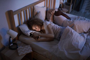 Yüksek açılı, kıvırcık saçlı genç bir kadının gece yatakta erkek arkadaşıyla yatarken akıllı telefon kullanmasının portresi.