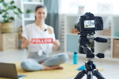 Evde egzersiz videosu kaydederken elinde FOLLOW işareti olan gülümseyen genç bir kadının bulanık portresi. Kameraya bakıyor, ön plana odaklanıyor, boşluğu kopyalıyor.