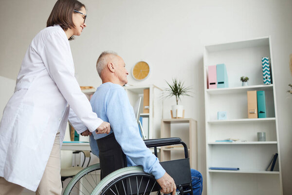 Side view portrait of smiling female doctor pushing senior man in wheelchair for examination or consultation in clinic, copy space