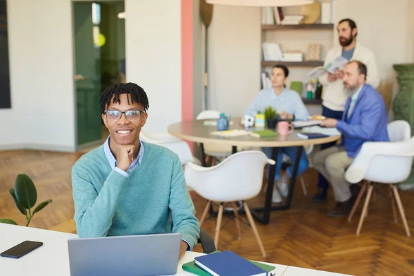 年轻的非洲裔美国人坐在写字台前 笔记本电脑上挂着微笑 他的同事们在他身后工作 — 图库照片