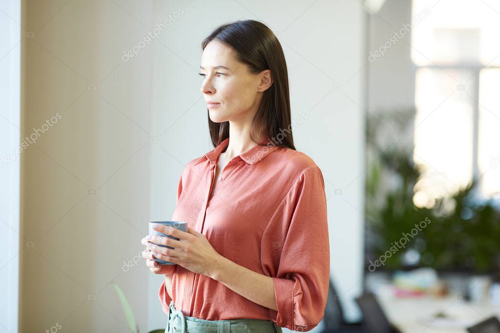 Young attractive woman wearing pink blouse standing in office room holding cup of coffee looking away