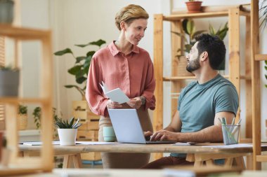 Modern ofis içinde masa başında ahşap detaylarla çalışan yakışıklı sakallı bir adamla konuşan kadın yöneticinin portresi.