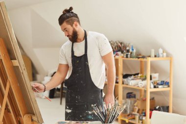 Geniş bir sanat stüdyosunda çalışırken sehpaya resim çizen çağdaş sakallı sanatçının belden yukarı portresi.
